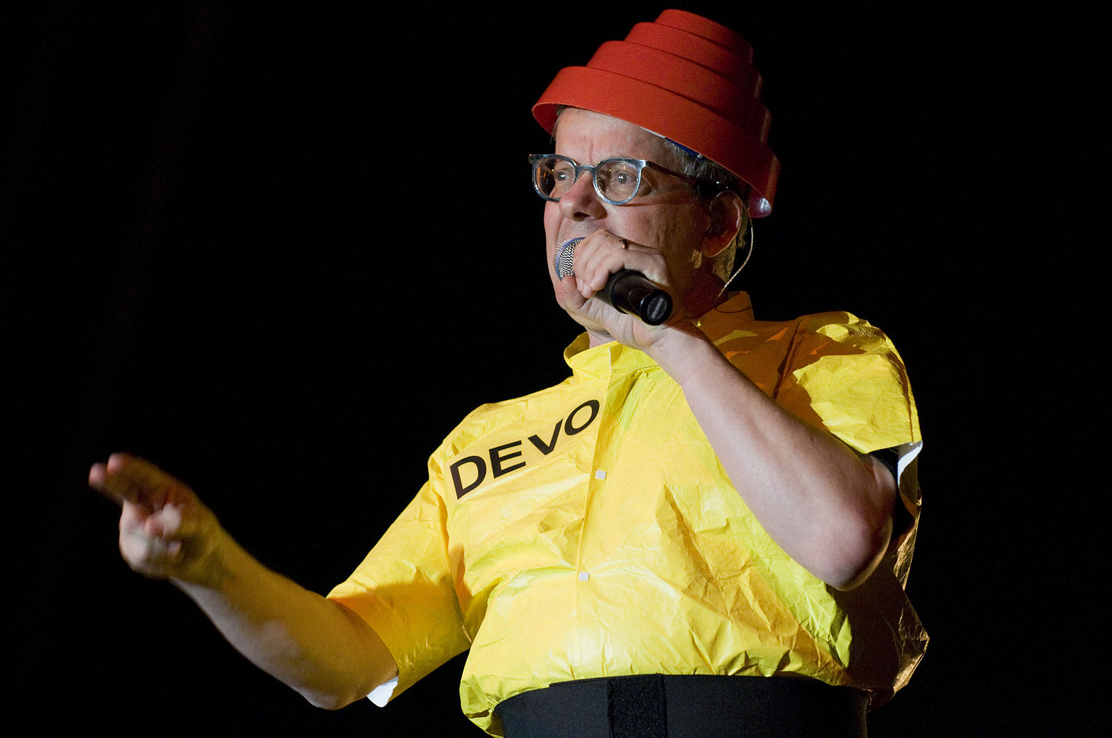 In a yellow shirt and red "power dome" helmet thing, Devo frontman Mark Mothersbaugh gestures while singing into a microphone.