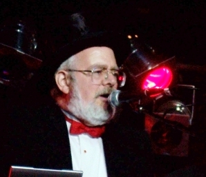Wearing a tuxedo with a red bowtie and a black top hat, Dr. Demento speaks to an audience... we can't tell, but he's probably saying something weird.