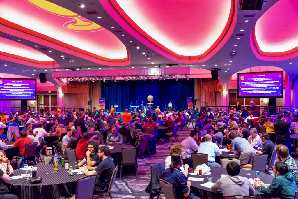 In a colorful ballroom, many attendees play trivia quizzes at round tables.